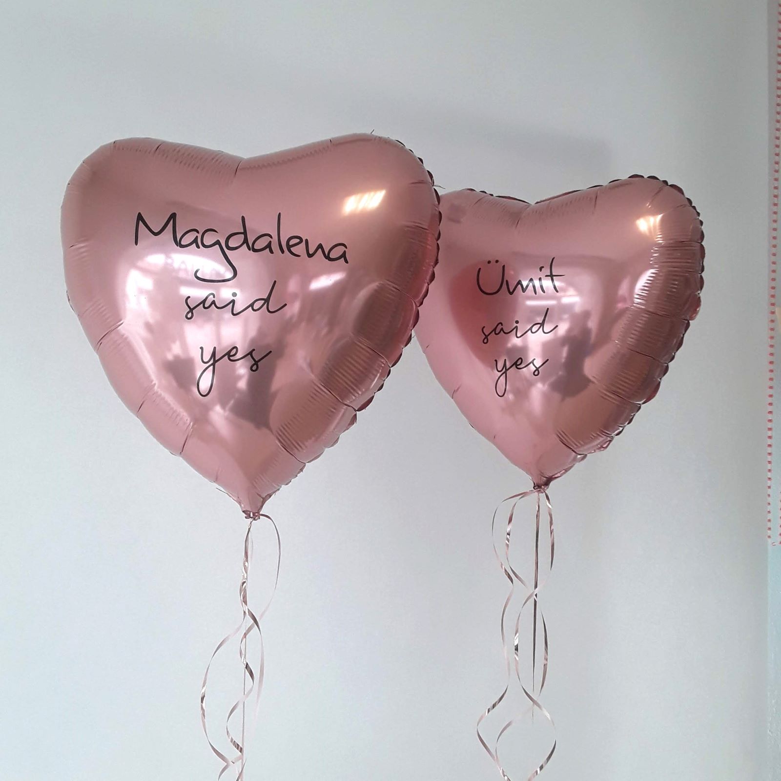 Zwei rosafarbene Folienballons in Herzform mit schwarzer Aufschrift „Magdalena said yes“ und „Ümit said yes“, jeweils mit passendem Geschenkband
